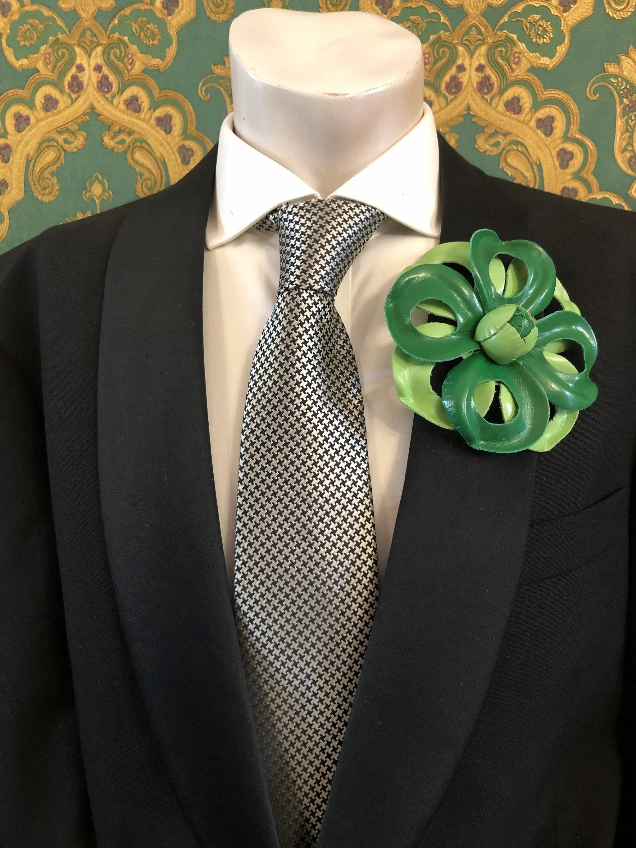 A Broochella Men's Viola del Pensiero handmade designer green leather flower-shaped brooch pin. It is displayed  on a black suit jacket, white dress shirt, and patterned grey tie on a mannequin torso The background is ornate green and gold patterned wallpaper.
