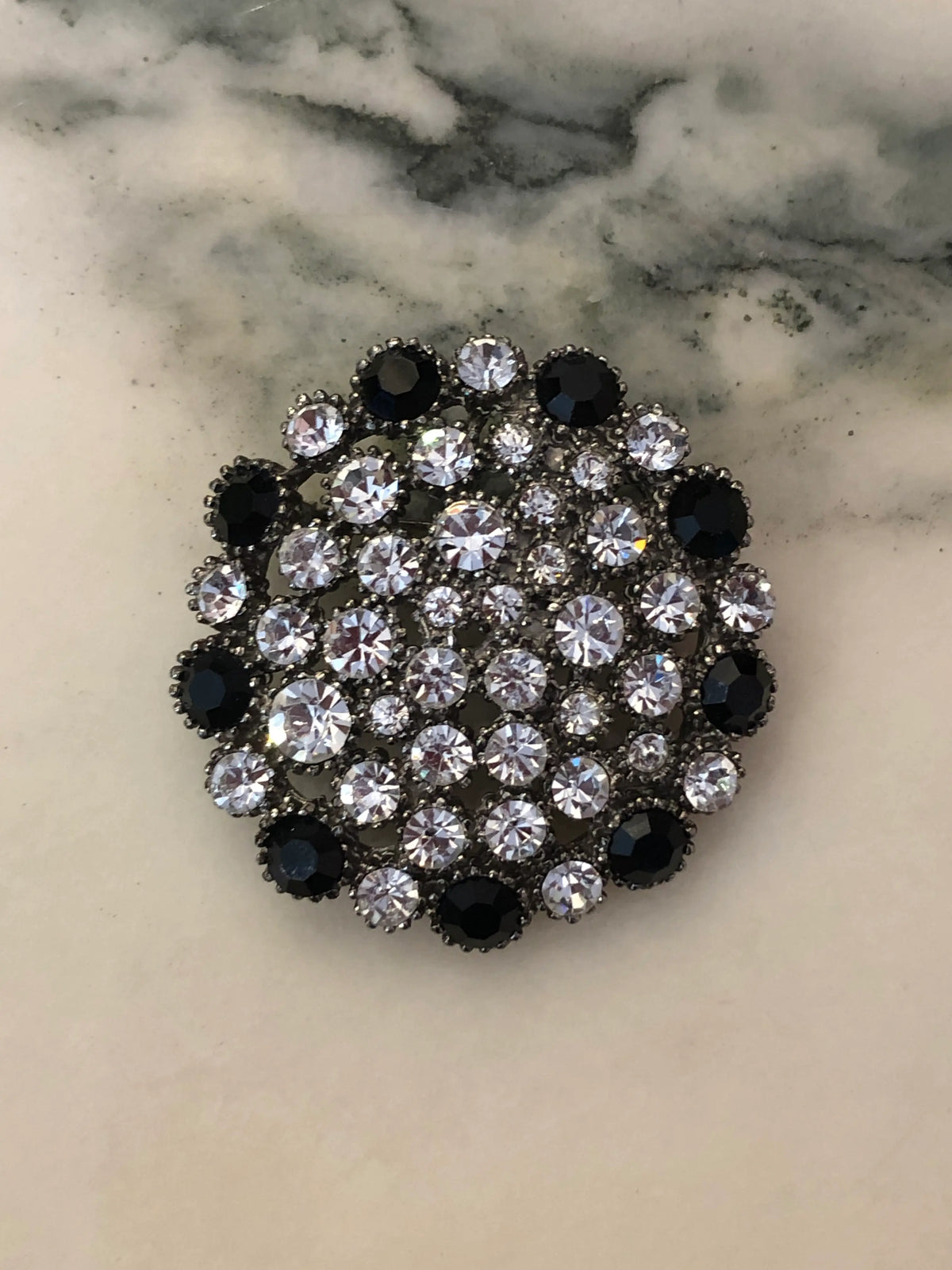 Broochella Men's Alessia Vintage Black & Crystal Brooch featuring clear crystals and jet-black stones in an ornate silver-tone circular setting on a cream and grey marble background.