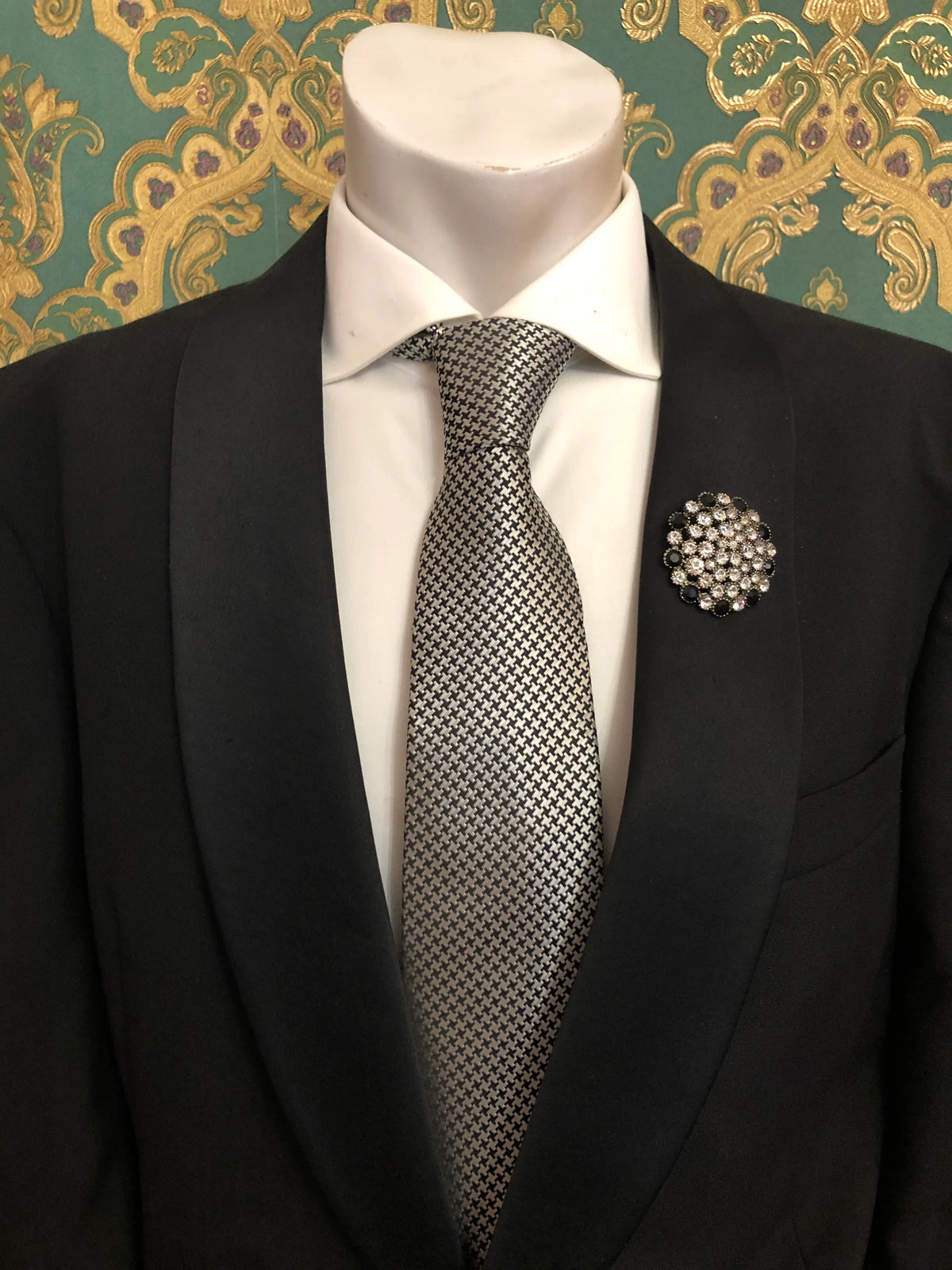 Broochella Men's Alessia vintage black and crystal brooch featuring a circular cluster of sparkling clear rhinestones surrounded by black stones, styled on the lapel of a black tailored suit with a white shirt and black-and-white patterned tie, against an ornate emerald green and gold damask wallpaper background.