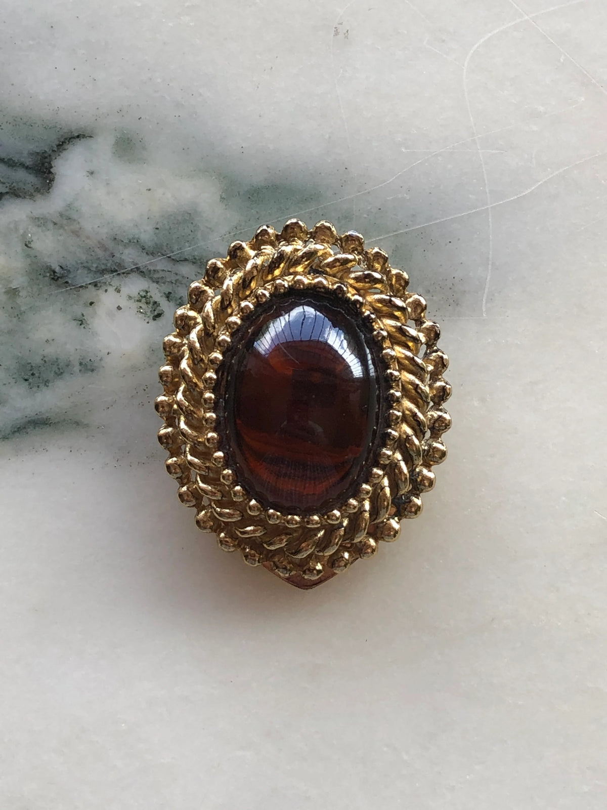 Broochella Men’s Amber Crest Vintage Tie Clip featuring a polished amber-brown oval cabochon framed by an ornate textured gold-tone setting, photographed on white marble with natural grey-green veining.