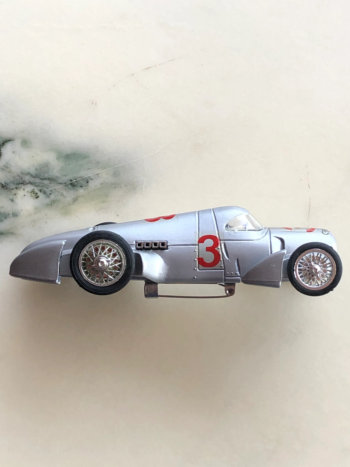 Broochella Men's Argento No.3 Vintage Formula One Racing Car Brooch photographed from above, showcasing its metallic silver body, red number 3 markings, aerodynamic rear fins, exposed wheels and enclosed cockpit.