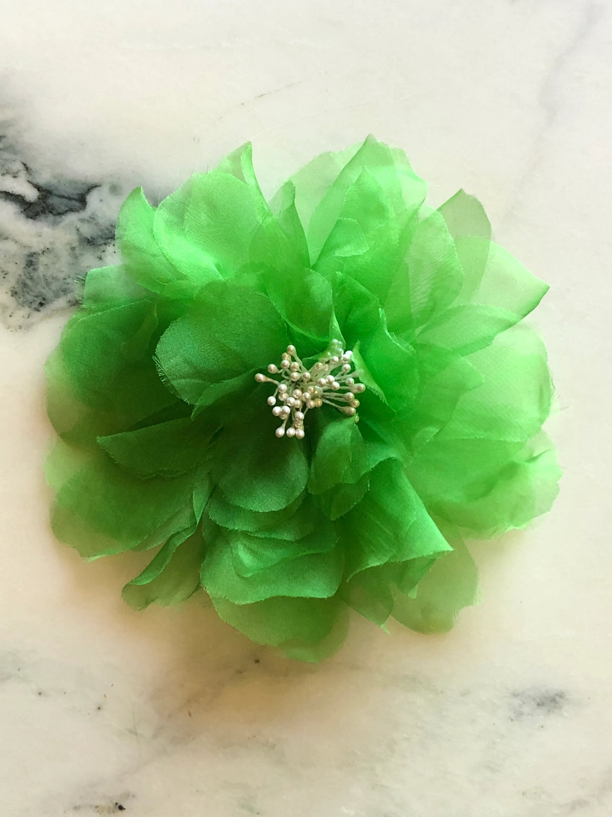 Broochella men’s Aubrieta handmade designer flower brooch in vibrant green fabric with pearl stamen detailing, photographed on a white and green veined marble background.