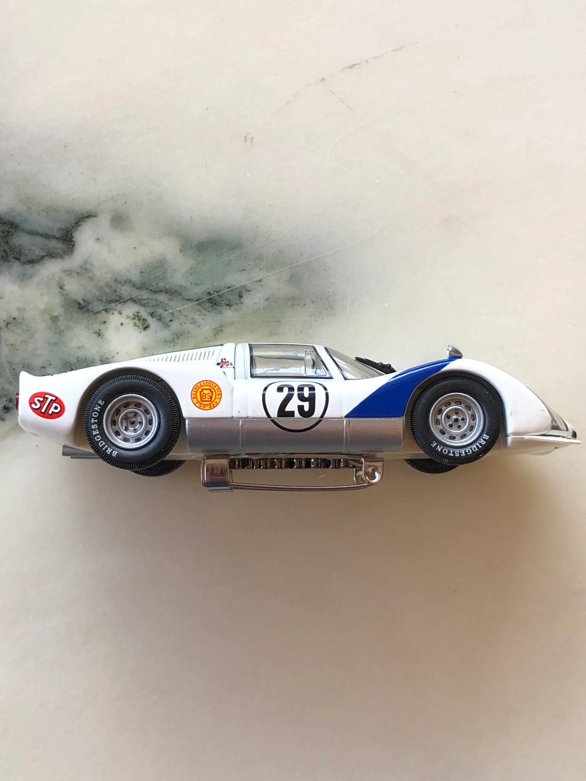 Broochella Men's Bianco Takumi No.29 Vintage Racing Car Brooch in side profile, featuring a white and blue racing design, number 29 decal, Bridgestone tyres, STP detailing, silver wheels and a visible metal brooch pin fastening.