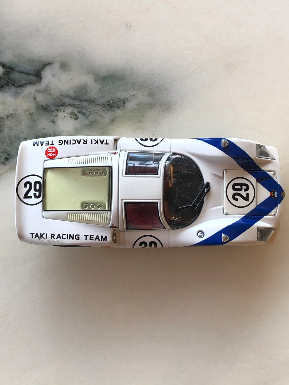Broochella Men's Bianco Takumi No.29 Vintage Racing Car Brooch photographed from above, showcasing white bodywork, blue racing stripes, number 29 markings, Taki Racing Team lettering and a clear rear engine cover.