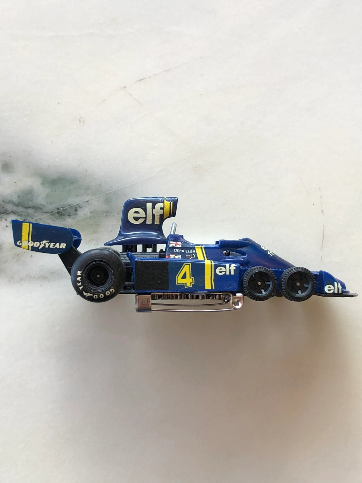 Broochella Men's Blu Tyrrell No.4 Vintage Formula One Racing Car Brooch in side profile, featuring six black racing tyres, yellow number 4, Elf and Goodyear logos, raised aerodynamic wings and a visible silver brooch pin fastening.