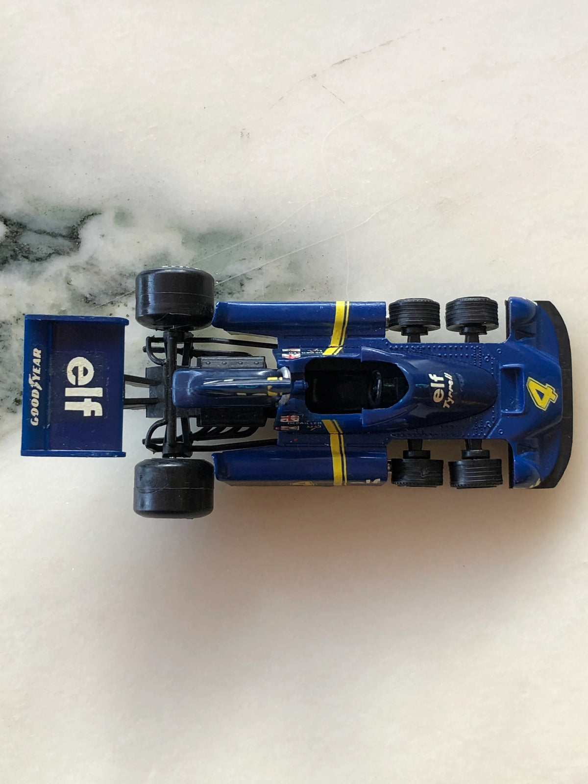 Broochella Men's Blu Tyrrell No.4 Vintage Formula One Racing Car Brooch photographed from above, showcasing its navy blue bodywork, yellow racing stripes, six-wheel design, open cockpit, Elf sponsorship and large rear wing.