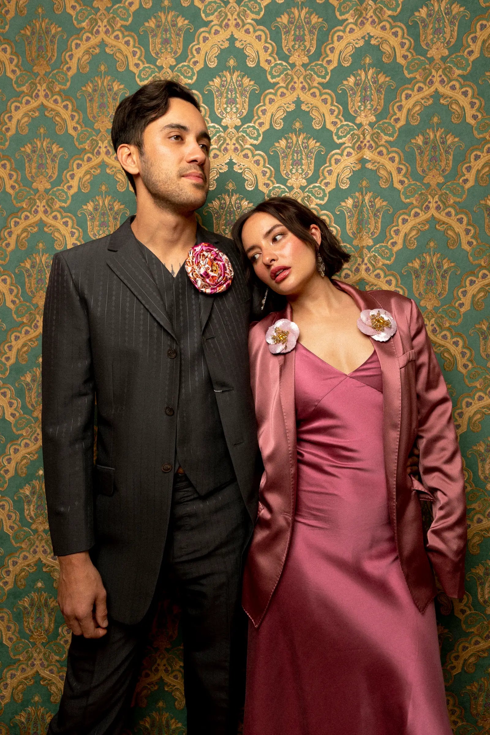 A man in a dark gray pinstripe suit with a pink floral Broochella Bougainville flower brooch in pink, stands next to a woman in a dusty rose satin dress and matching jacket, both wearing matching floral lapel pins, posed against a green and gold damask wallpaper background.