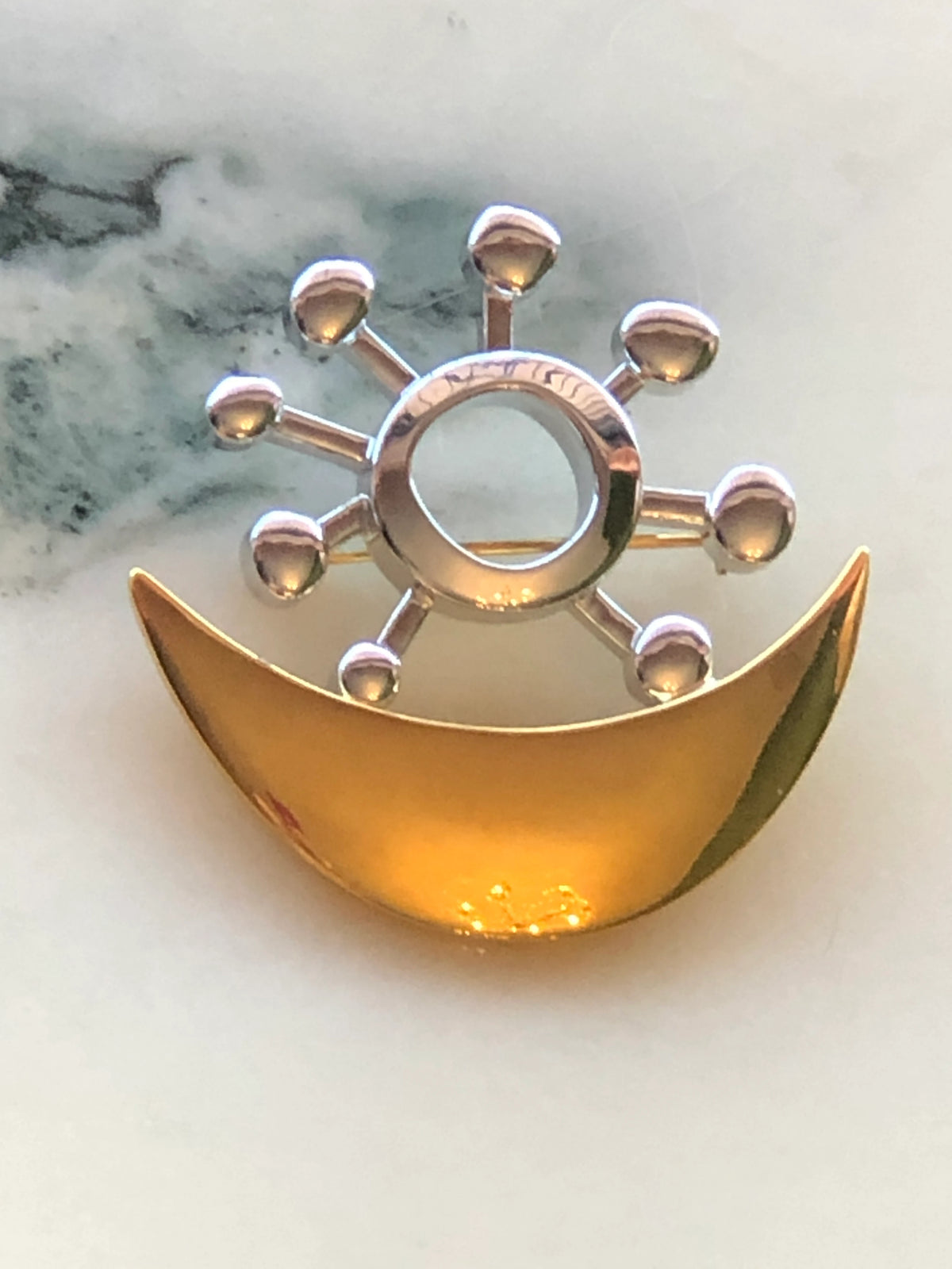 Broochella Men’s Brooches with Meaning Love and Harmony Brooch featuring an abstract polished gold crescent with a sculptural silver-tone circular motif, photographed on a pale white marble background with grey veining.