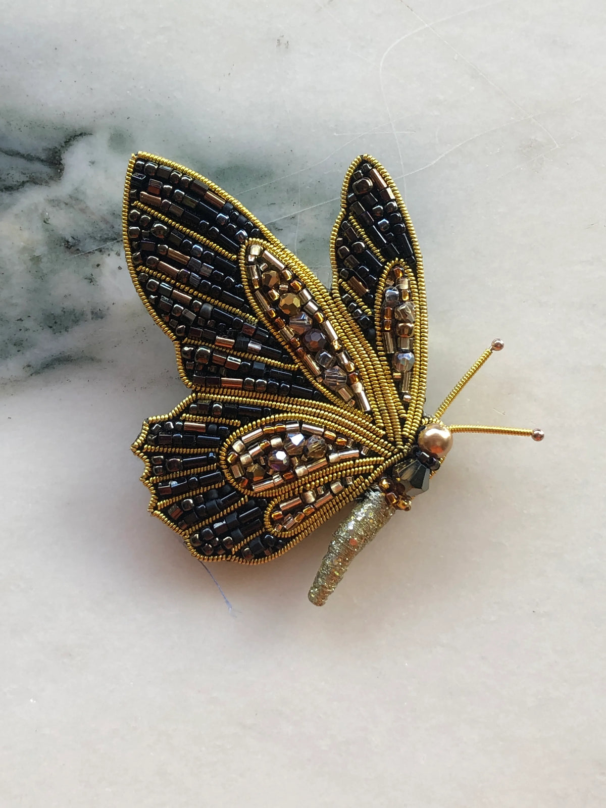 Broochella Men’s Butterfly Brooch featuring black, bronze and gold beadwork with crystal embellishments and metallic detailing, photographed on a white marble background with grey-green veining.