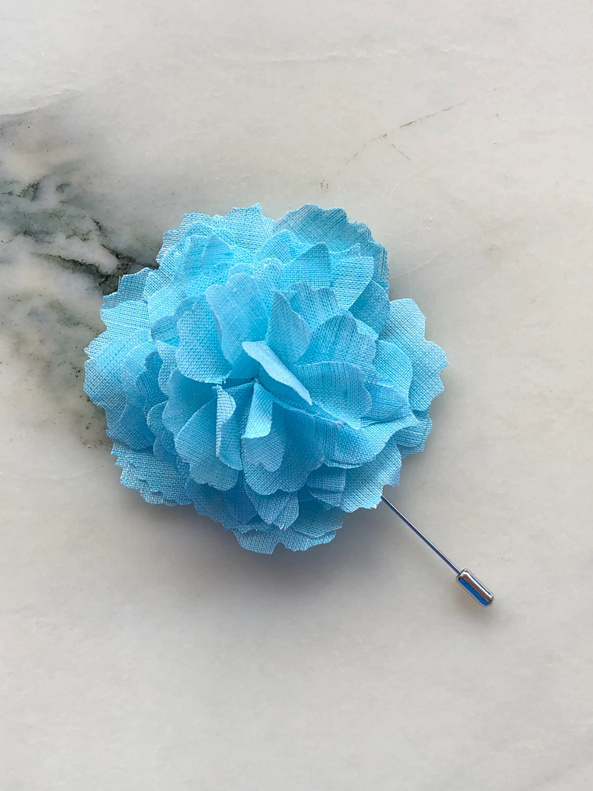 Broochella Men’s Carnation Flower Brooch in vibrant aqua fabric with layered petals and silver stick pin, photographed on a white marble background with grey-green veining.