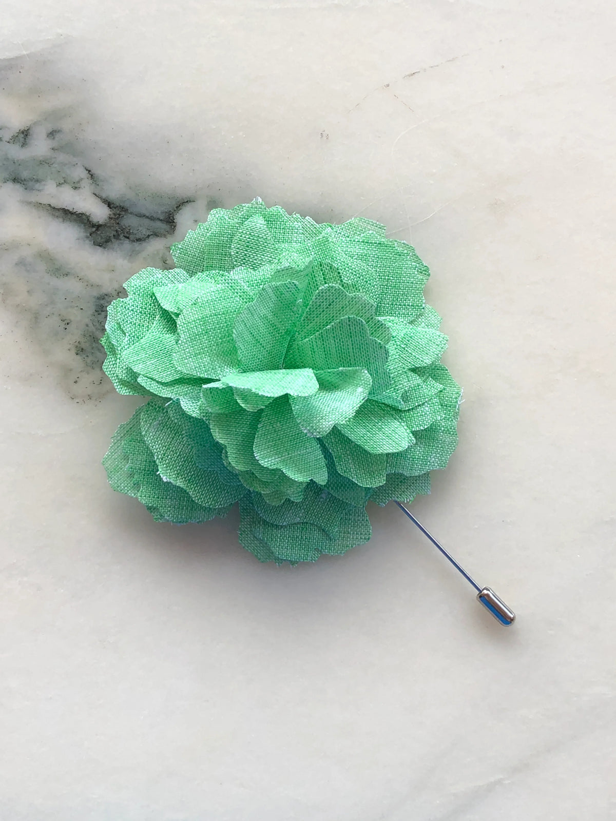 Broochella Men’s Carnation Flower Brooch in fresh green fabric with layered petals and silver stick pin, photographed on a white marble background with grey-green veining.