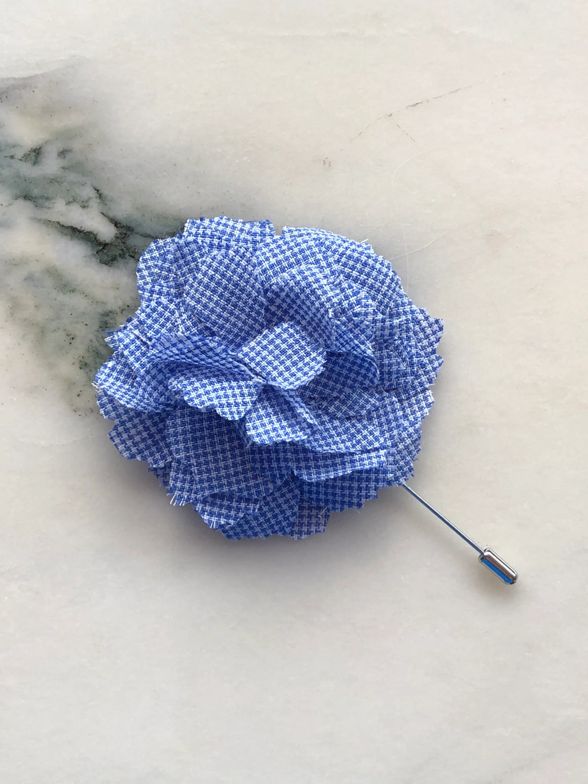 Broochella Men’s Carnation Flower Brooch in navy patterned fabric with layered petals and silver stick pin, photographed on a white marble background with grey-green veining.