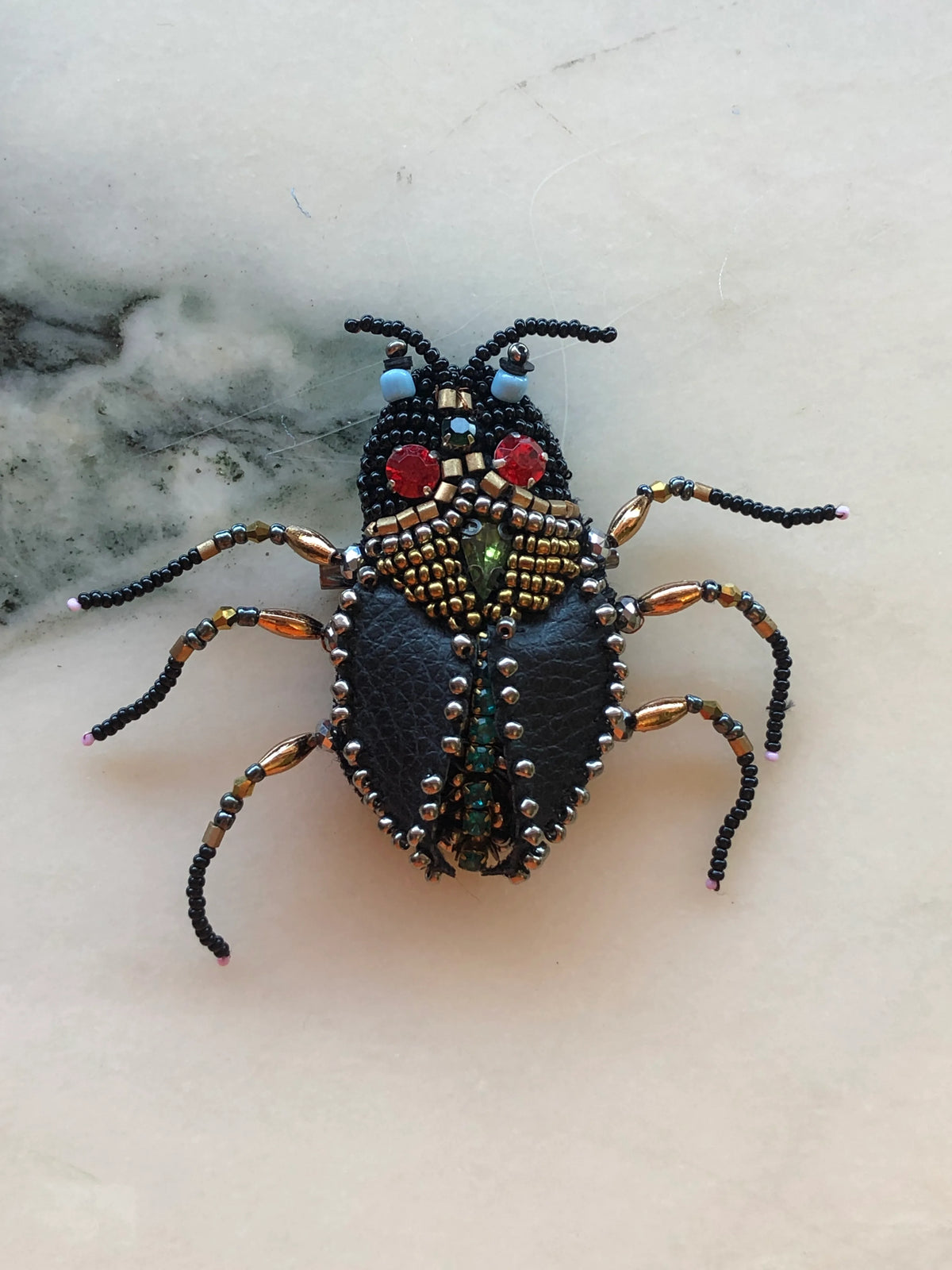 Broochella Men’s Cockroach Brooch featuring black leather wings, intricate black and gold beadwork, red crystal eyes and articulated beaded legs, photographed on a white marble background with grey-green veining.