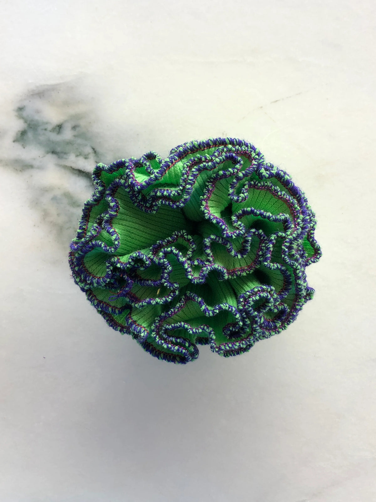 Broochella Men’s Corallo Brooch in vivid green fabric with contrasting purple and green ruffled edging, photographed on a white marble background with grey-green veining.