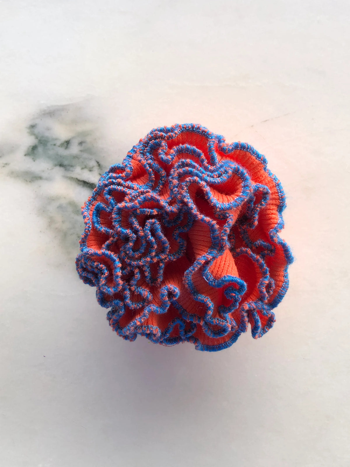 Broochella Men’s Corallo Brooch in bright orange fabric with contrasting blue ruffled edging, photographed on a white marble background with grey-green veining.
