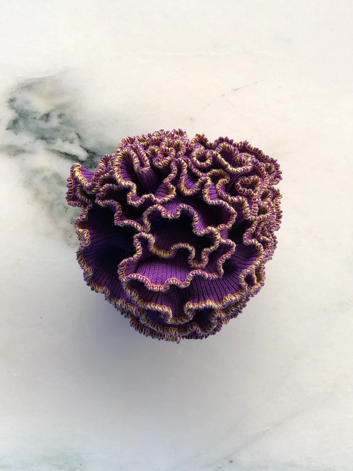 Broochella Men’s Corallo Brooch in rich purple fabric with contrasting golden yellow ruffled edging, photographed on a white marble background with grey-green veining.