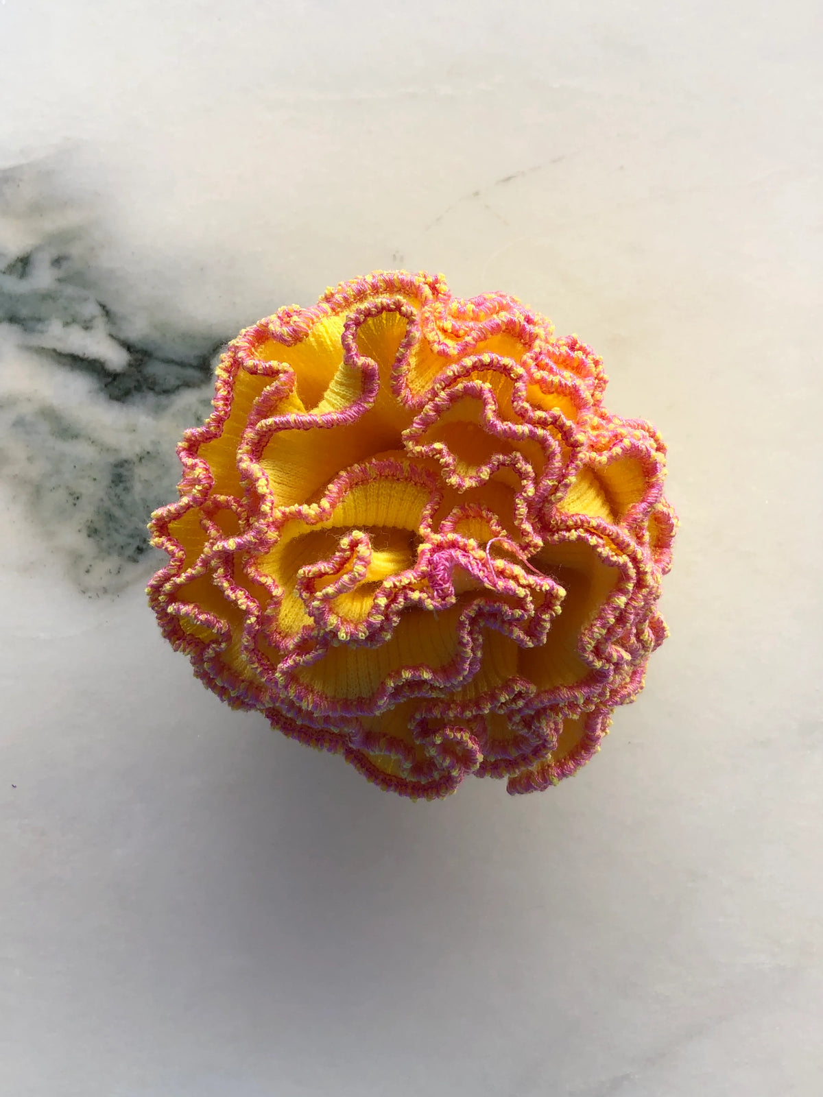 Broochella Men’s Corallo Brooch in bright yellow fabric with contrasting pink and yellow ruffled edging, photographed on a white marble background with grey-green veining.