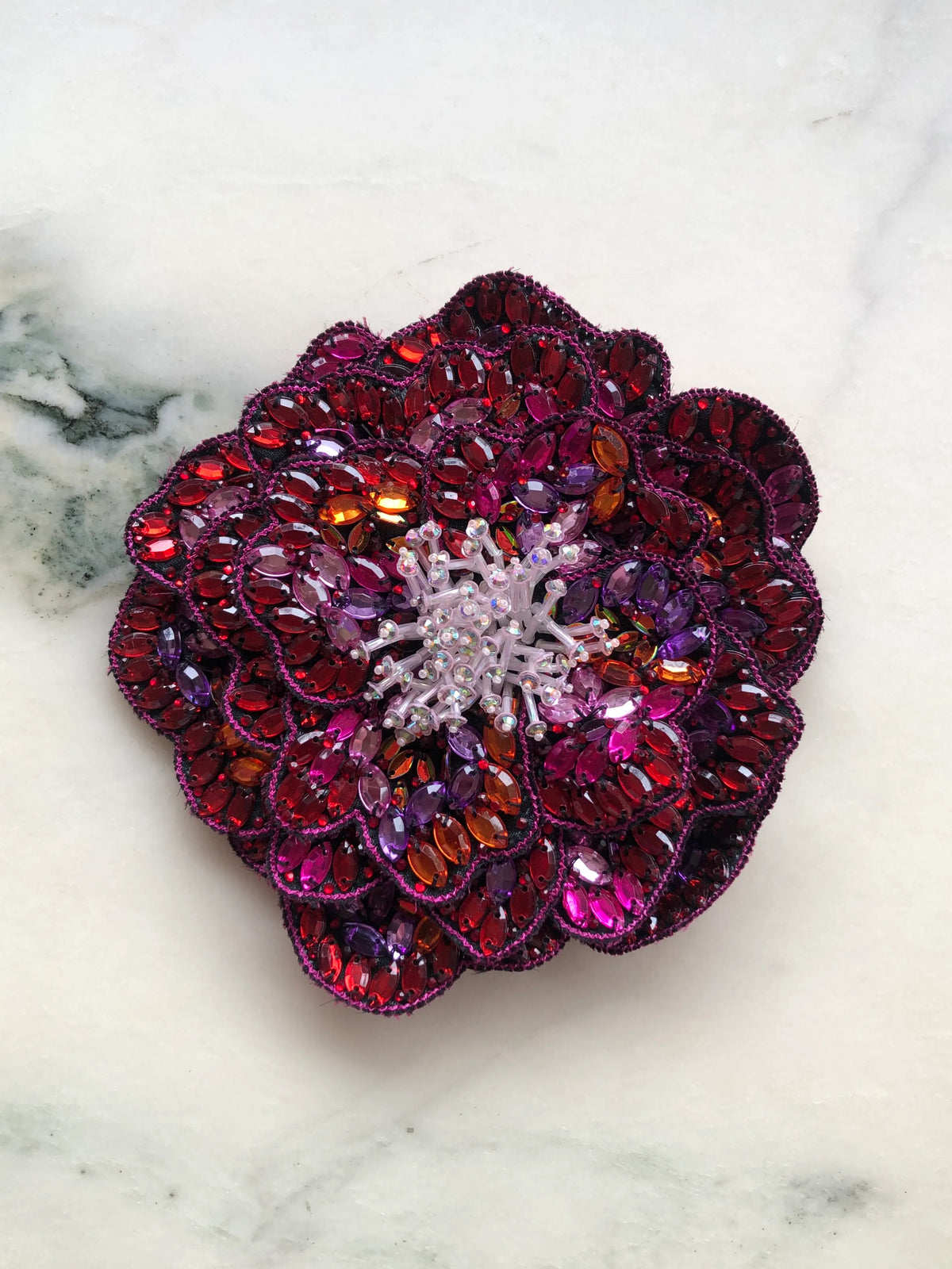 Broochella Men's Cristallino Hand-Beaded Brooch Pin, featuring a sculptural flower with layered petals embellished with sparkling red, burgundy, fuchsia, purple and orange crystals, finished with delicate iridescent beading at the centre. A statement floral brooch for men's blazers, jackets and formalwear, photographed on white marble with grey-green veining.