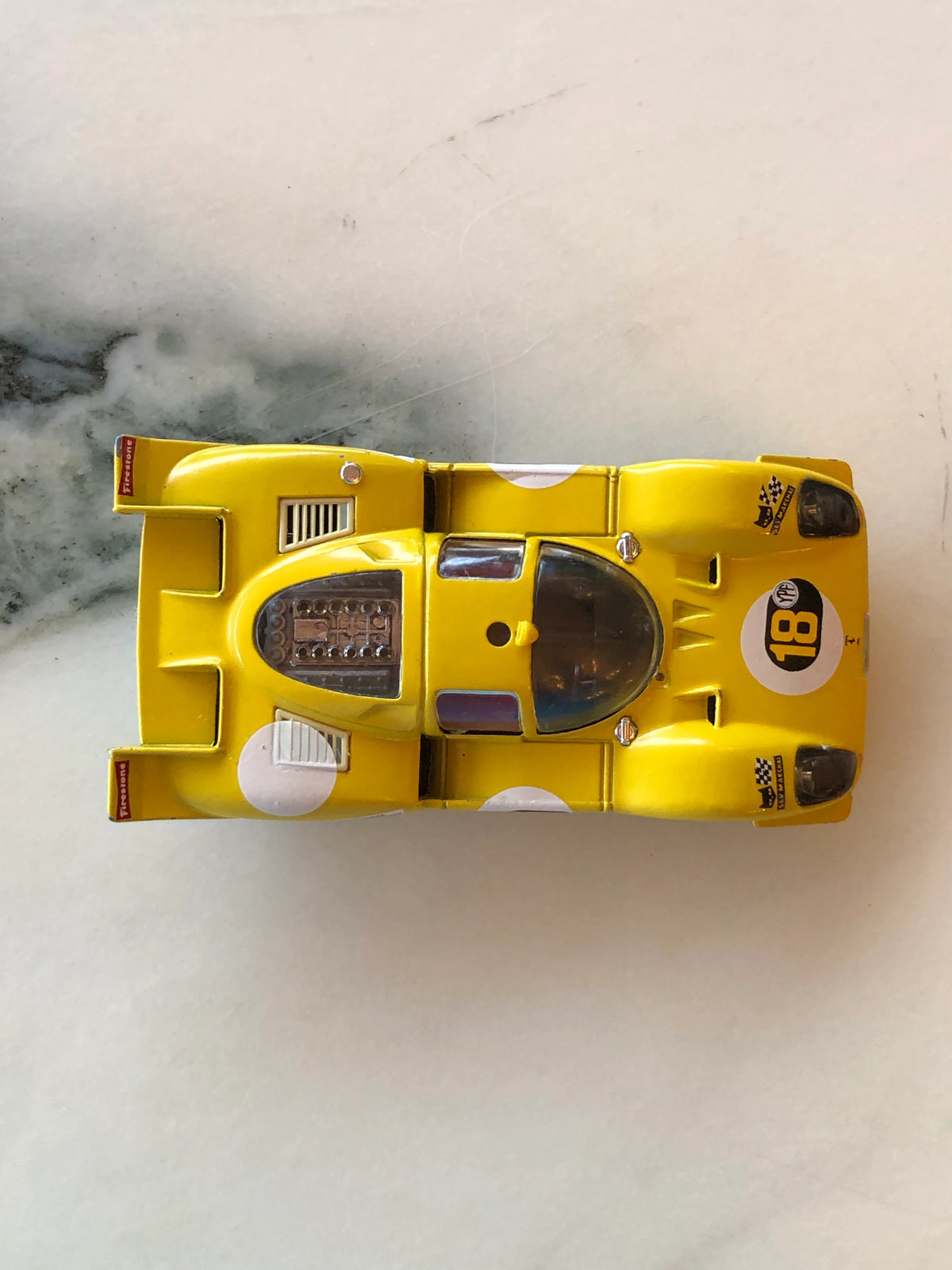 Broochella Men's Giallo Modena No.18 Vintage Racing Car Brooch photographed from above, showcasing its yellow body, number 18 racing decal, transparent cockpit, exposed engine detailing and wide rear spoiler.