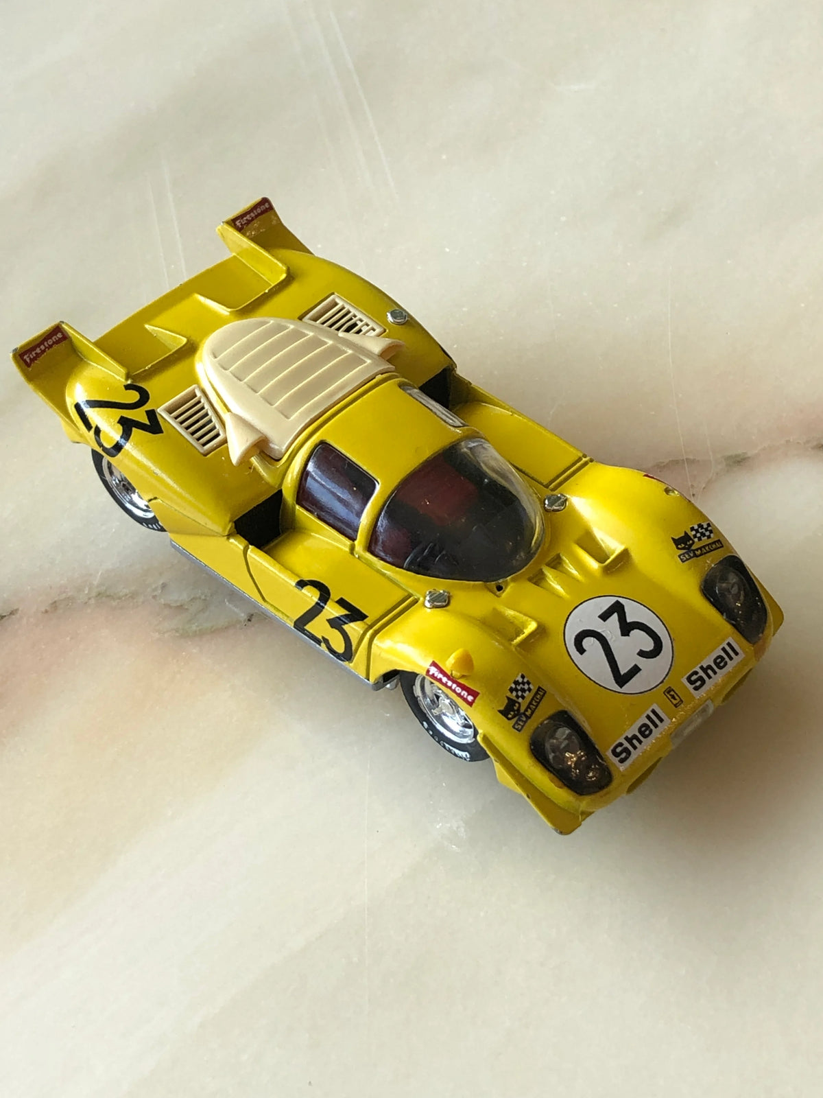 Broochella Men's Giallo Sprint No.23 Vintage Endurance Racing Car Brooch photographed from an elevated angle, featuring a bright yellow racing car with black number 23 decals, aerodynamic rear wing, clear cockpit canopy, cream engine cover and vintage Shell and Firestone racing details.