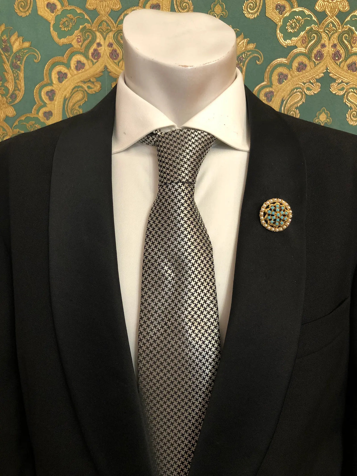 Broochella Men's Ginevra vintage turquoise and pearl brooch featuring a circular gold-tone design with vibrant turquoise-blue stones, delicate pearl accents and intricate lattice detailing, styled on the lapel of a black tailored suit with a crisp white shirt and black-and-white houndstooth tie, against ornate emerald green and gold damask wallpaper.