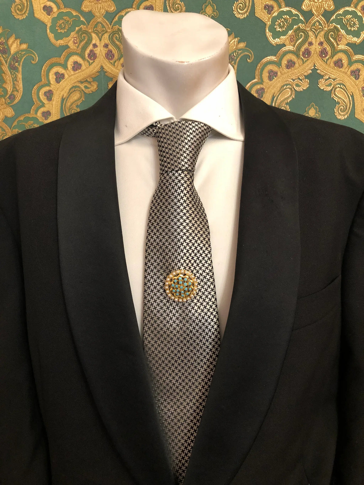 Broochella Men's Ginevra vintage gold-tone brooch featuring a circular cluster of turquoise-blue stones framed by luminous pearl-like beads, pinned to a black-and-white houndstooth necktie with a white shirt and black tailored suit, photographed against decorative emerald green and gold damask wallpaper.
