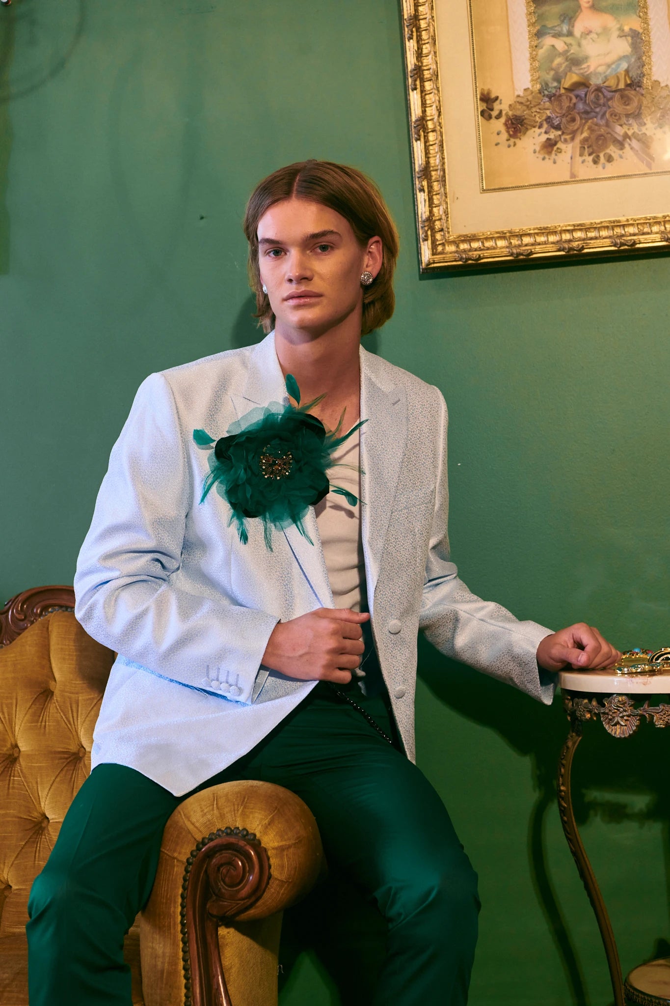 A man wearing a light blue blazer with a Broochella la Duchessa large green feathered flower brooch on the chest, paired with dark green trousers, seated in a yellow tufted armchair against a dark green wall.