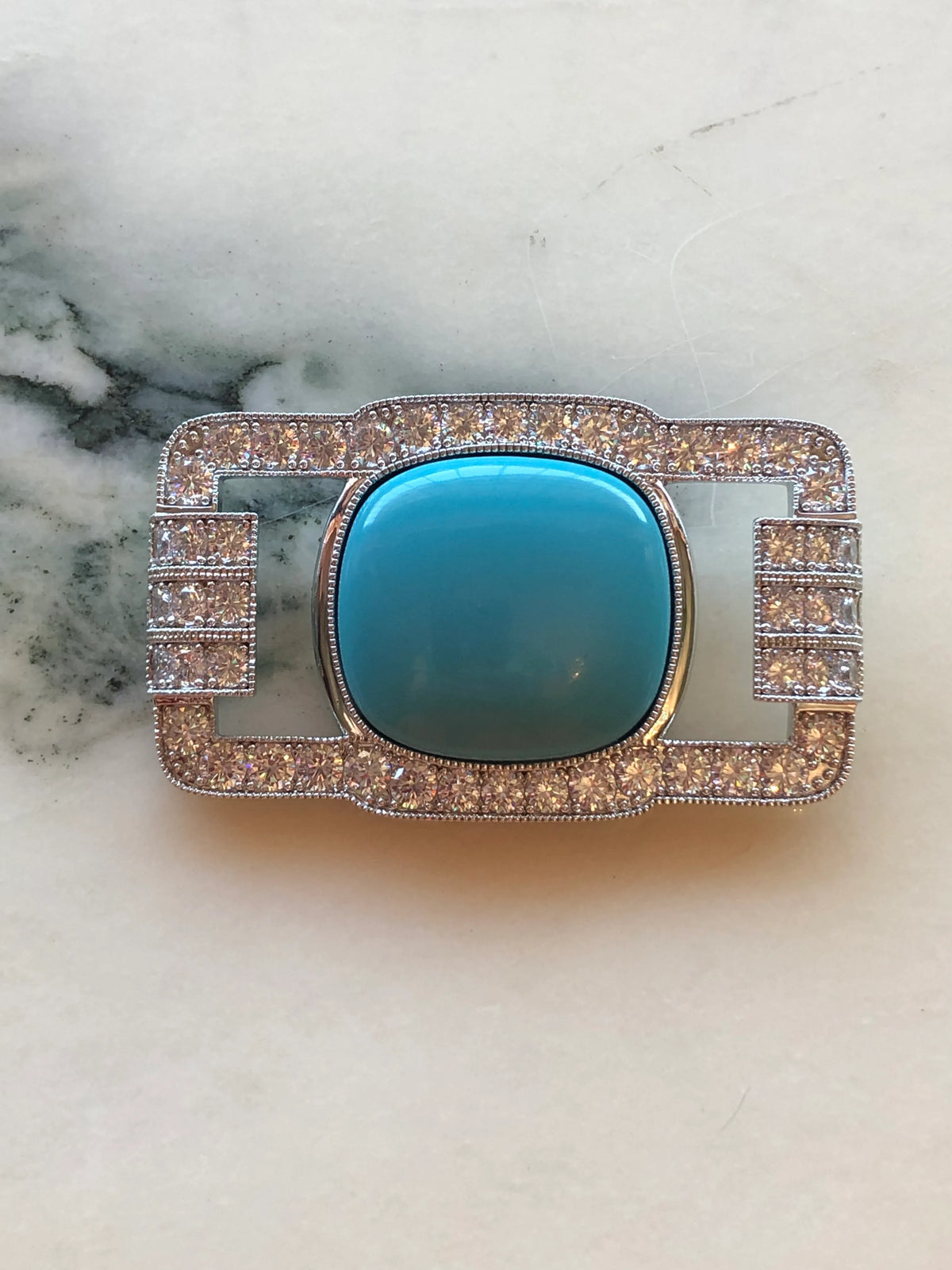 Broochella Men’s Libertenè Brooch featuring a polished turquoise blue centre stone framed by sparkling clear crystals in a silver-tone geometric setting, photographed on a pale white marble background with grey veining.