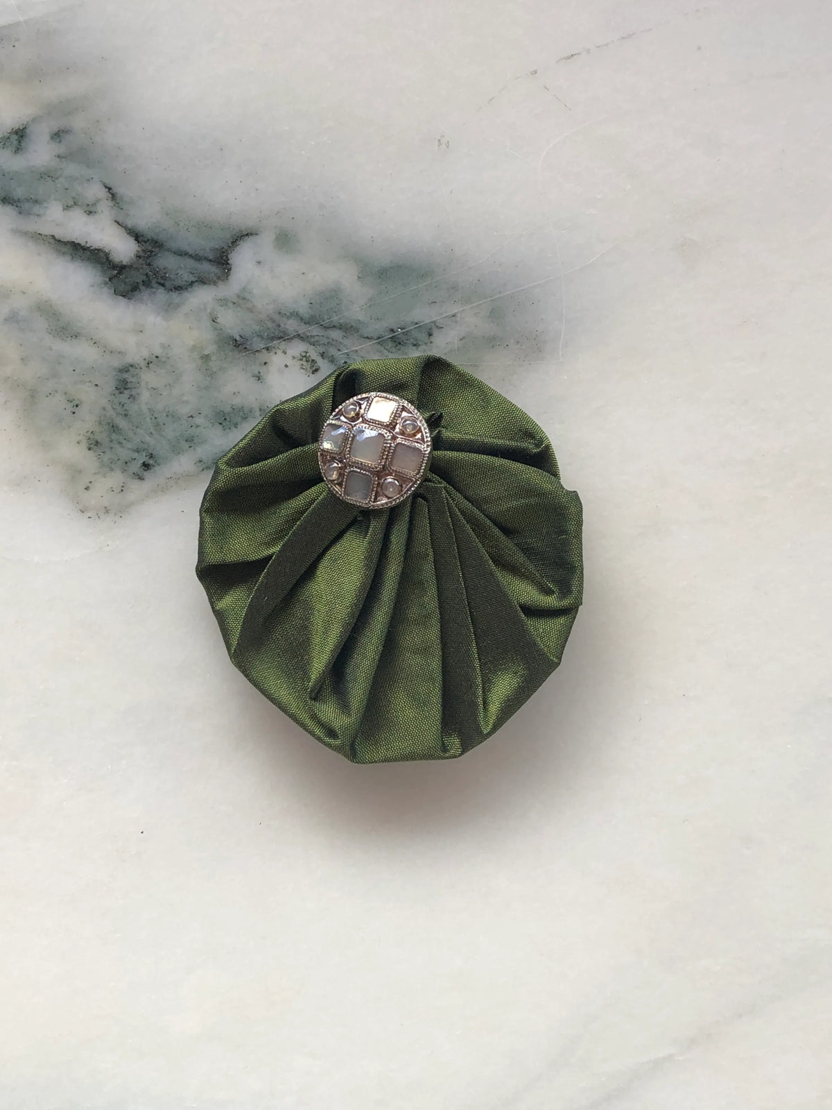 Broochella Men’s Lollipop Brooch in olive green, small size, featuring gathered silk fabric with a decorative silver-tone centre, photographed on a pale white marble background with grey-green veining.