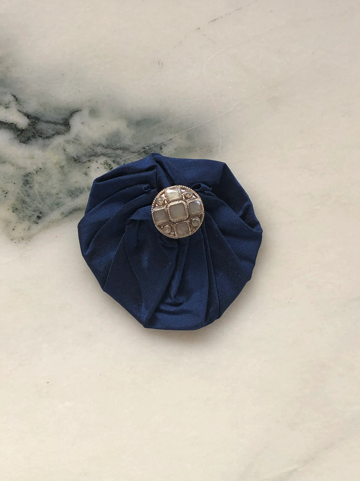Broochella Men’s Lollipop Brooch in navy, small size, featuring gathered silk fabric with a decorative silver-tone centre, photographed on a pale white marble background with grey-green veining.
