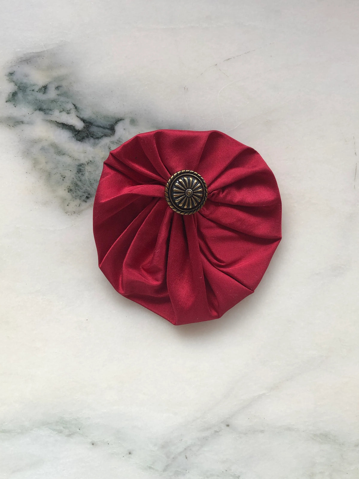 Broochella Men’s Lollipop Brooch in red, medium size, featuring gathered silk fabric with an antique-style metal centre, photographed on a pale white marble background with grey-green veining.