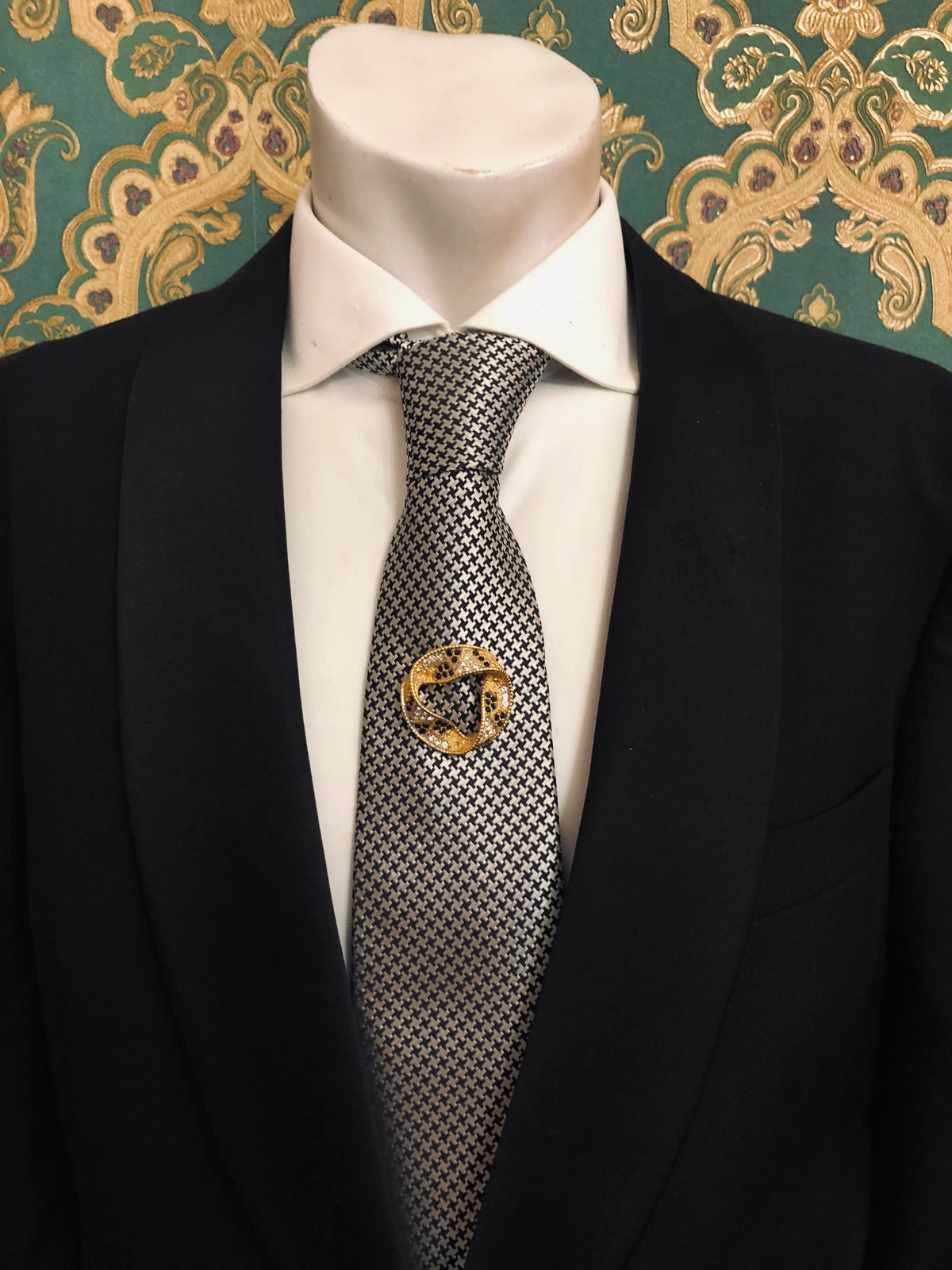 Broochella Men's Ottavia vintage black and crystal swirl brooch featuring a sculptural gold-tone circular design with sparkling clear rhinestones and contrasting black stones, pinned to a black-and-white houndstooth patterned tie with a crisp white shirt and black tailored suit, photographed against emerald green and gold damask wallpaper.