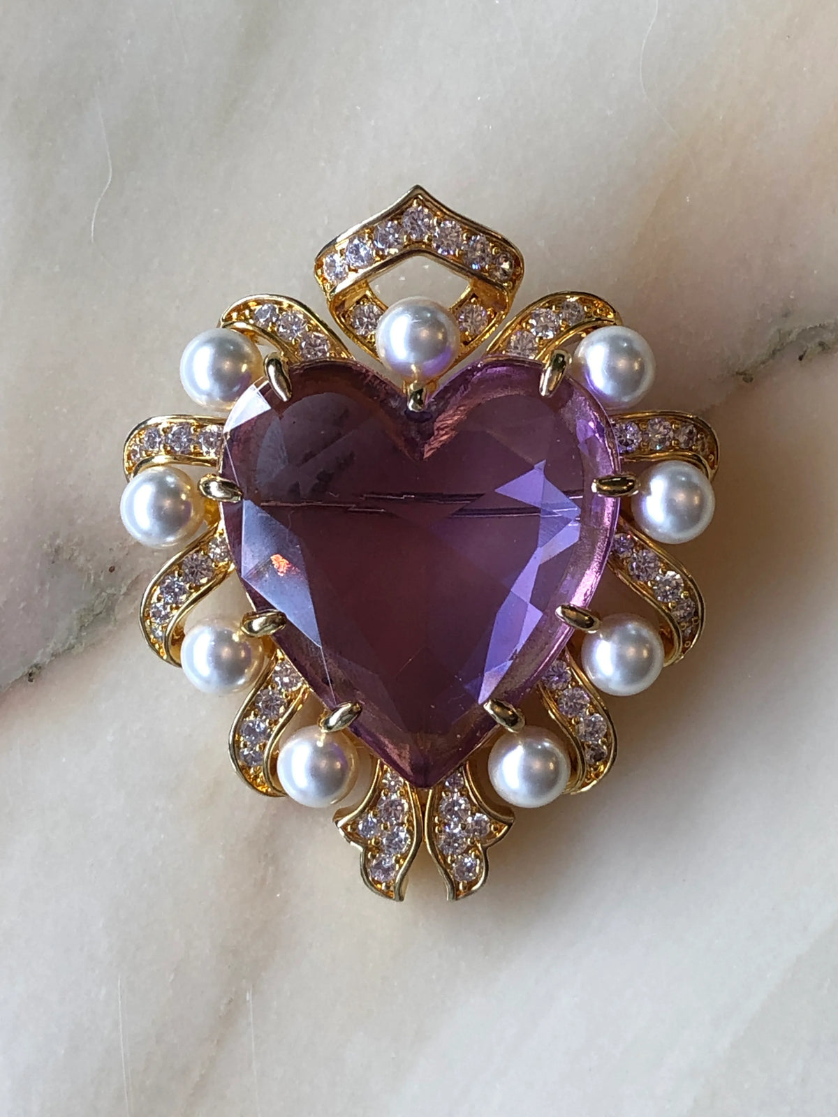 Broochella Men’s Romeo & Juliet Brooch featuring a faceted purple heart framed in gold, white pearls and sparkling crystals, photographed on a pale marble background with subtle grey veining.