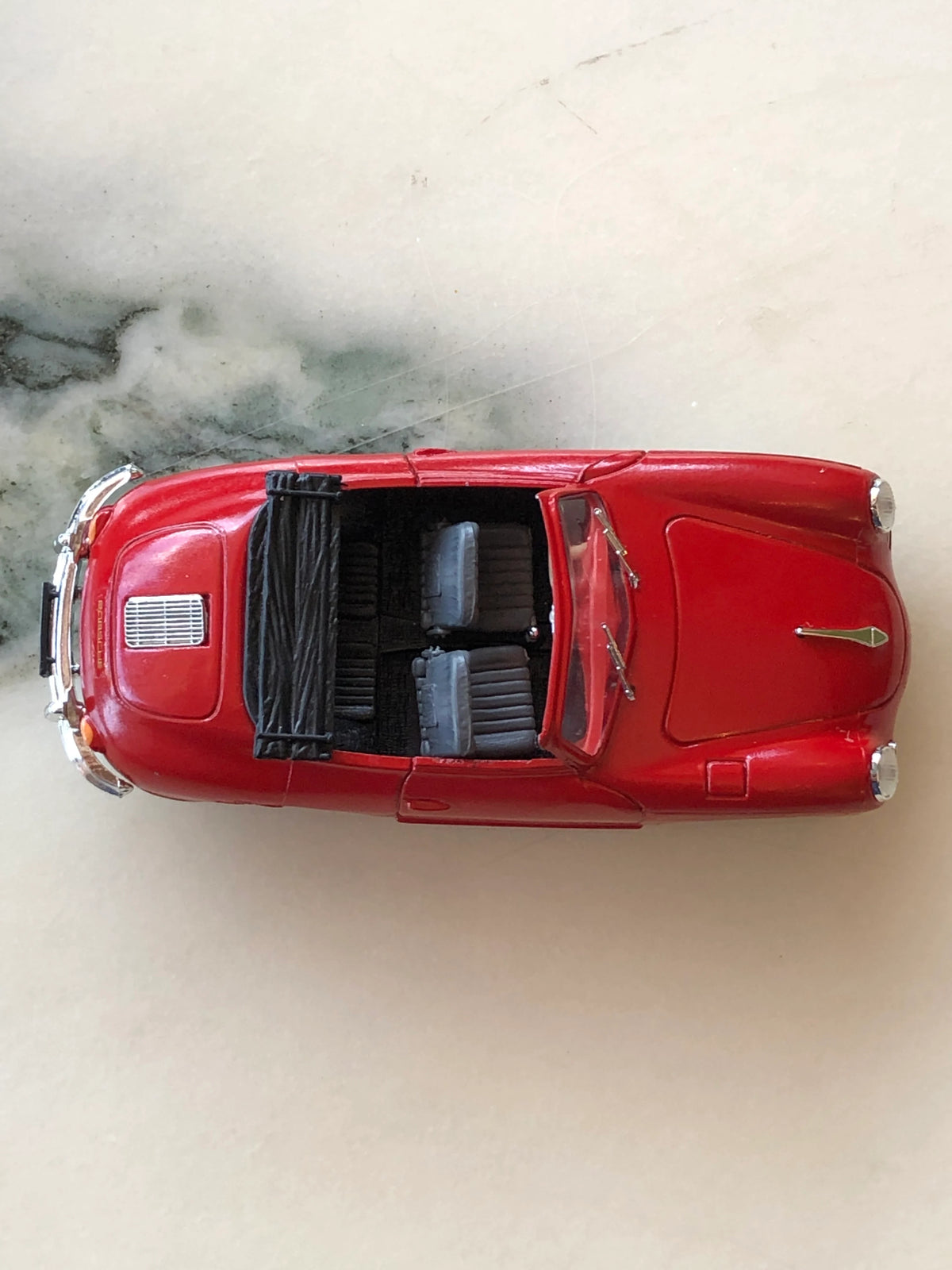 Broochella Men's Rosso Cabrio No.356 Vintage Convertible Car Brooch photographed from above, showcasing its red bodywork, open cockpit, two dark seats, folded black convertible roof and chrome windscreen detailing.