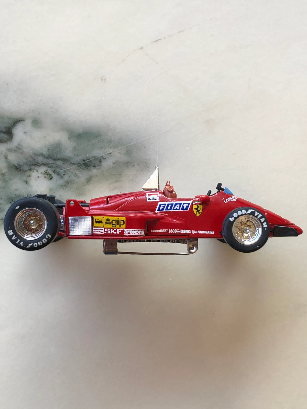 Broochella Men's Rosso Maranello No.27 Vintage Ferrari Formula One Racing Car Brooch in side profile, featuring a red racing body, Ferrari shield, FIAT and Agip sponsor decals, gold-tone wheels and silver brooch pin fastening underneath.