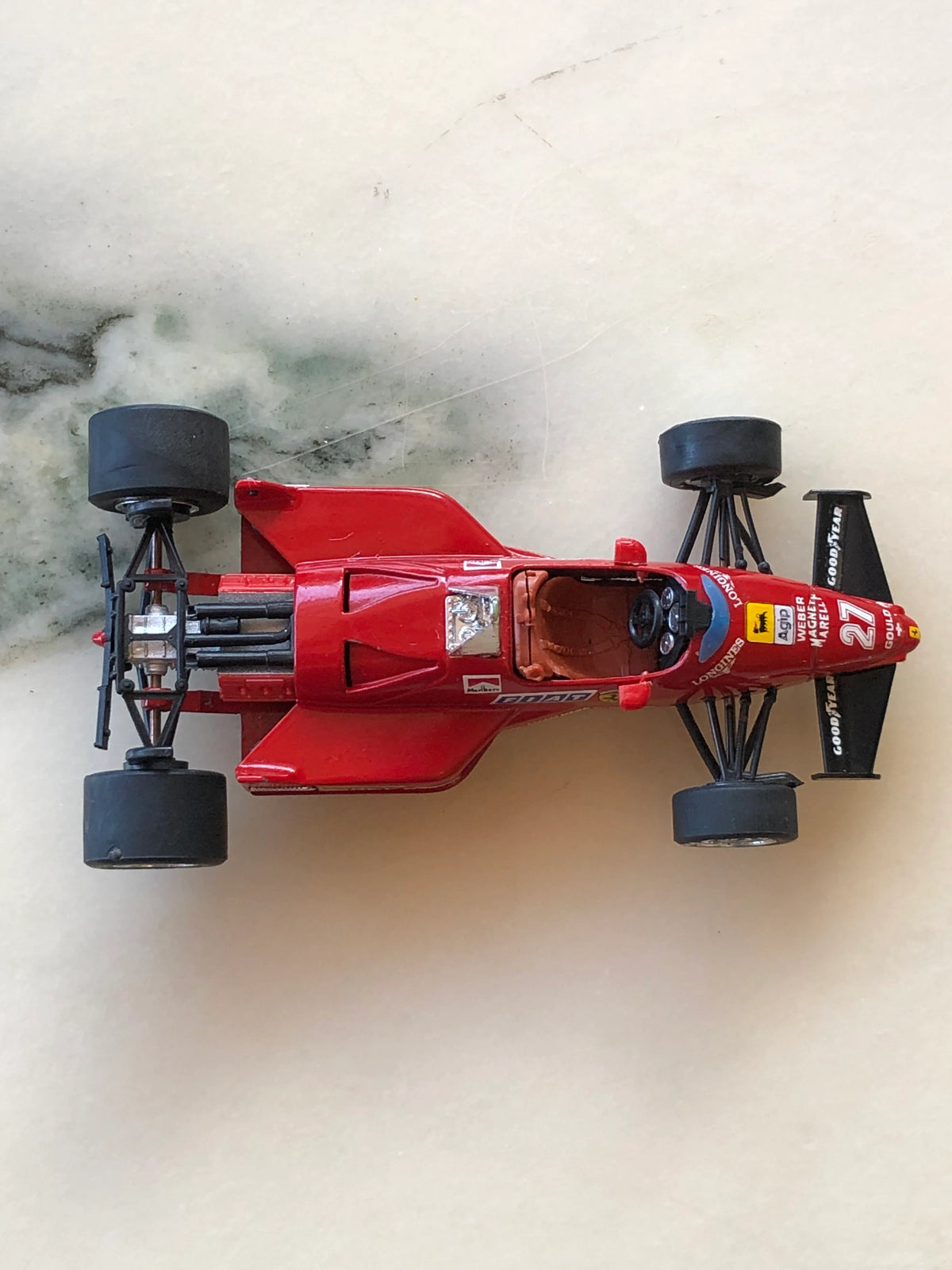 Broochella Men's Rosso Maranello No.27 Ferrari Formula One Racing Car Brooch viewed from above, showcasing its red aerodynamic body, number 27 racing decals, brown cockpit interior, exposed rear engine and black Goodyear tyres.
