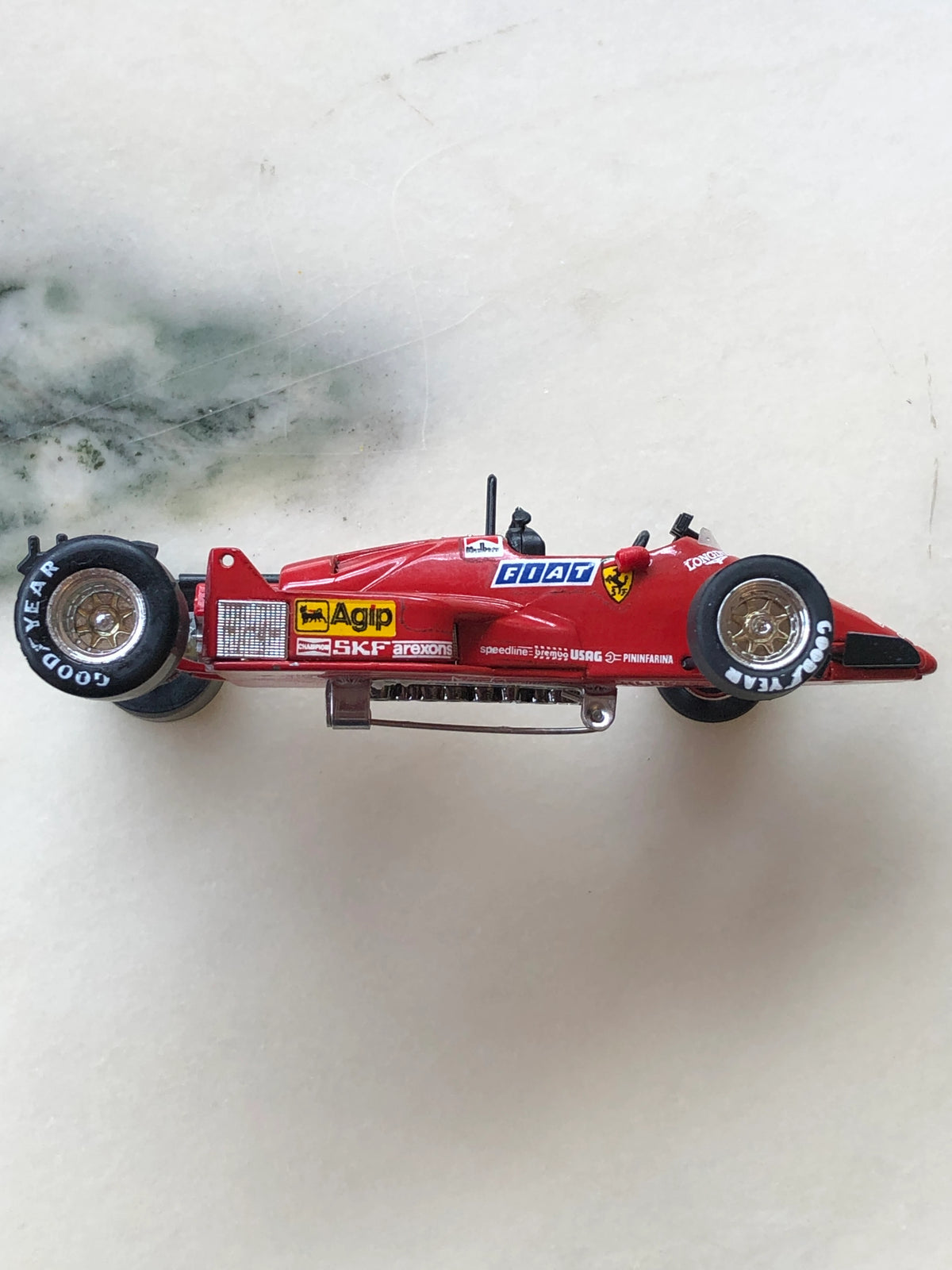 Broochella Men's Rosso Monza No.28 Ferrari Formula One Racing Car Brooch in side profile, featuring a red vintage Formula One car with gold wheel rims, black Goodyear tyres, Ferrari shield, FIAT and Agip racing decals and a silver brooch pin fastening underneath.