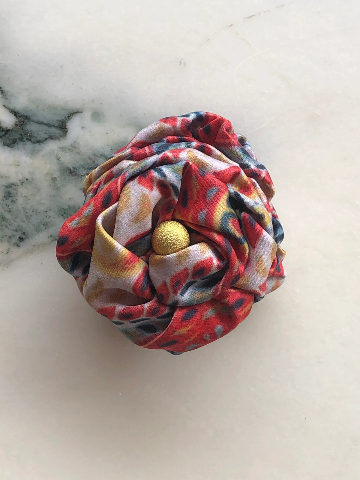Broochella Men’s Sartorial Bloom Brooch, a handmade designer fabric flower with sculptural folded petals in red, blue, gold and soft pink print, finished with a gold centre and photographed on white marble.