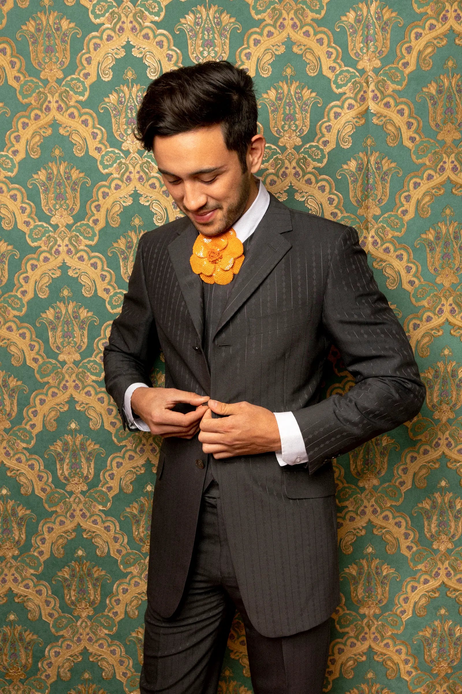 A man wearing a dark gray pinstripe three-piece suit with a white dress shirt and a Broochella Men's Scintillante bright orange flower-shaped necktie, standing against a green and gold damask wallpaper background.