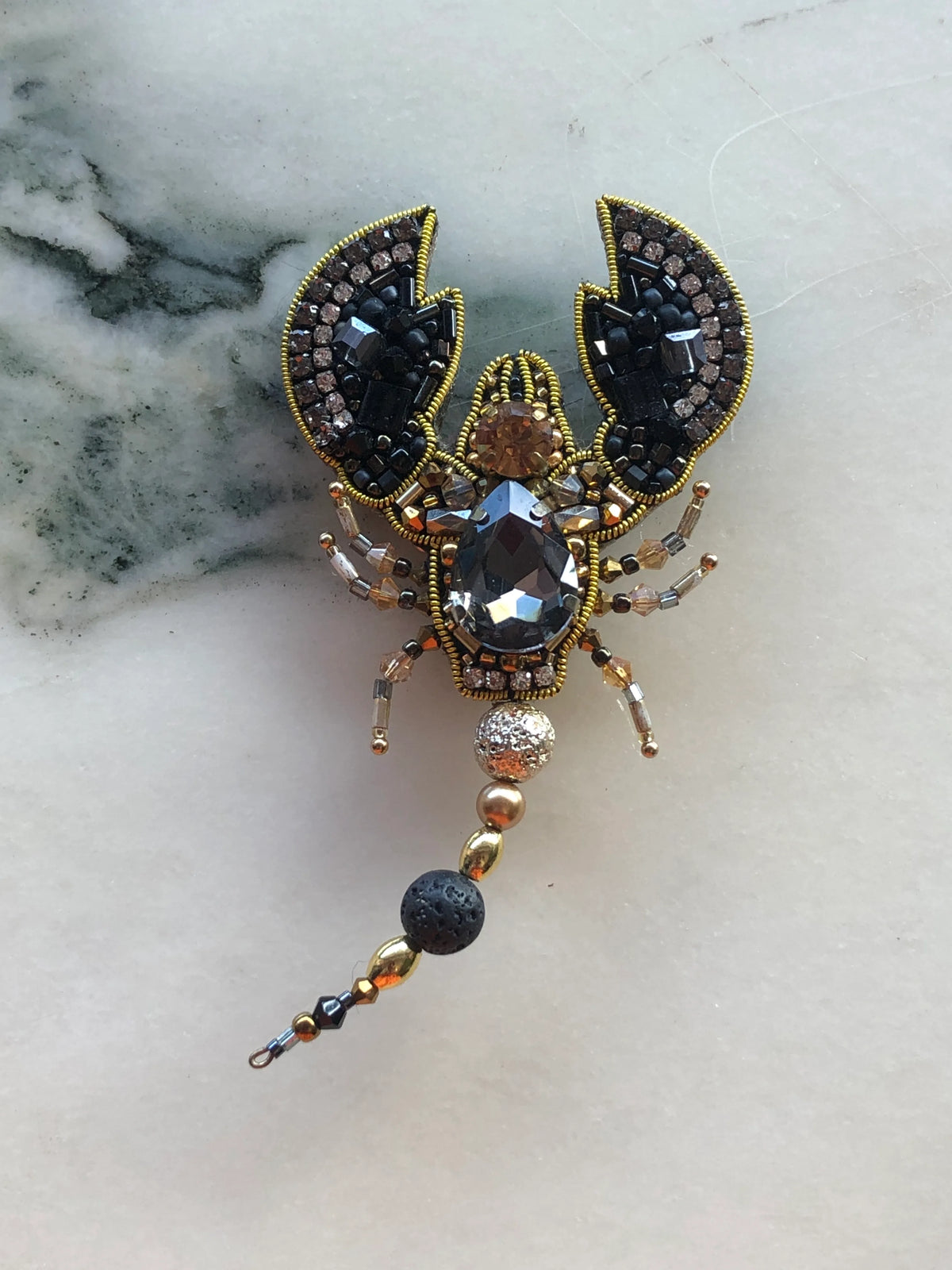 Broochella Men’s Scorpion Brooch featuring black and smoky crystal beadwork, gold detailing and a beaded articulated tail, photographed on a white marble background with grey-green veining.
