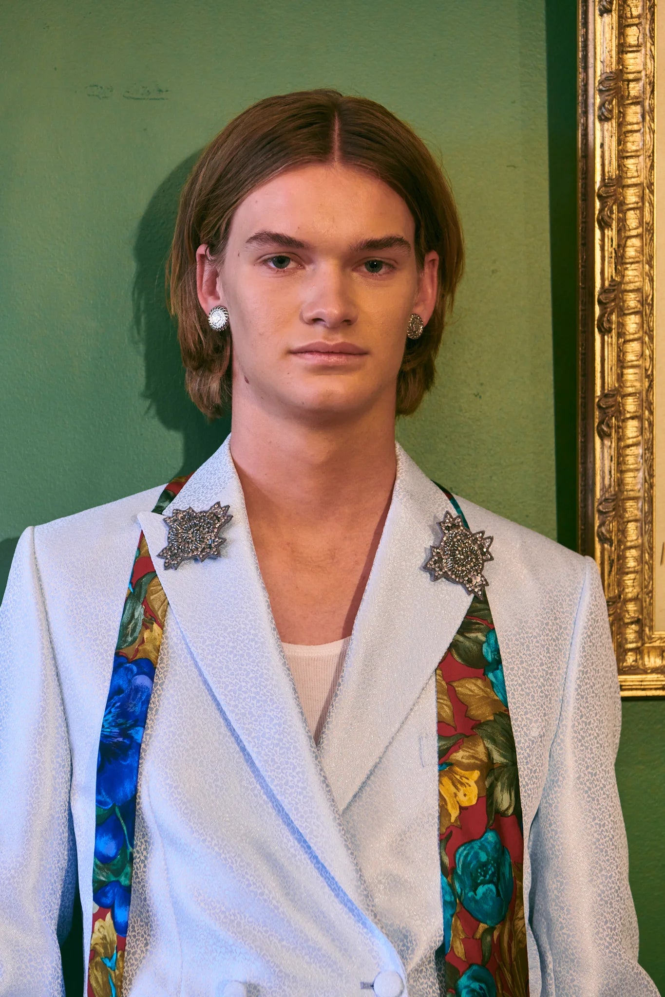 A male model with shoulder-length light brown hair stands against a green wall wearing a white textured blazer with ornate Broochella Stella Men's silver brooch and a colourful floral patterned scarf draped over his shoulders.