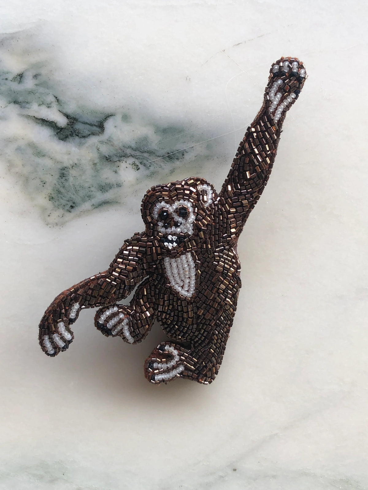 Broochella Men’s Tropicana Monkey Brooch Pin, a playful statement monkey design intricately hand-beaded in bronze, brown, black and white beads, photographed on white marble with soft grey-green veining.