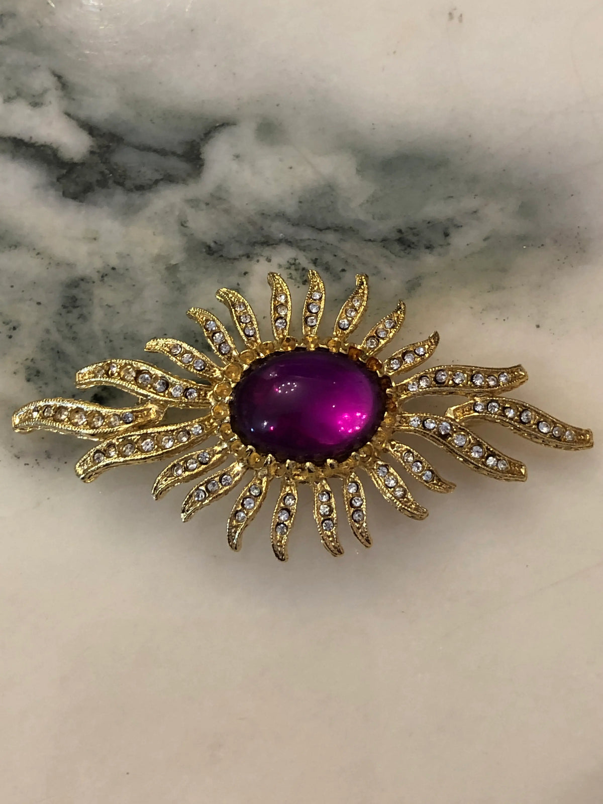 Broochella Men's Valentina Vintage Purple Sunburst Brooch featuring a deep-purple oval cabochon surrounded by gold-tone crystal-set rays on a cream and grey marble background.