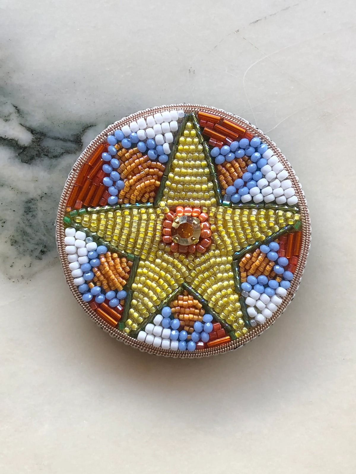 Broochella Men’s Viva La Stella Brooch Large, hand-beaded with a golden star and colourful blue, orange and white beadwork, photographed on a white marble background.