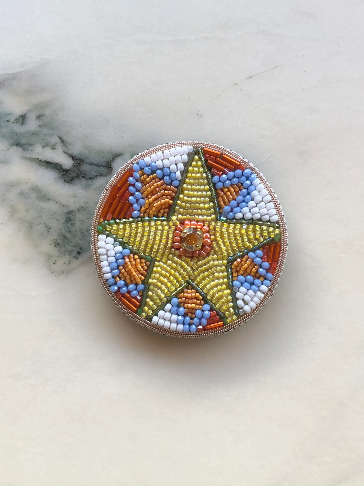 Broochella Men’s Viva La Stella Brooch Small, hand-beaded with a golden star and colourful blue, orange and white beadwork, photographed on a white marble background.
