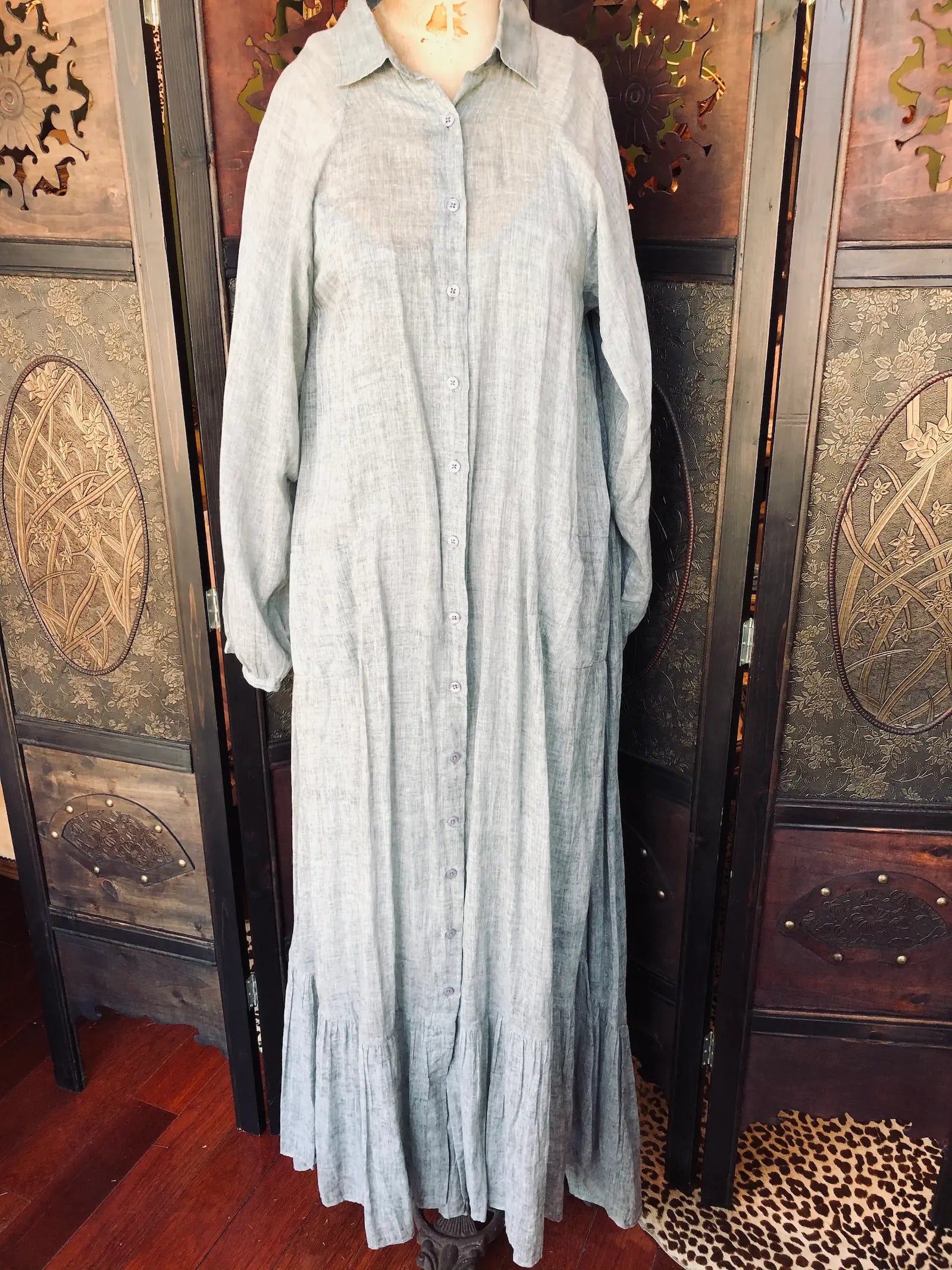Broochella- Mes Demoiselles Paris Size:36, 100% Cotton, Soft muted green-grey tone, Button-through front, Long sleeves, Maxi length dress. It is pictured on a vintage mannequin. The background is an ornate, vintage, dark wood room divider. 