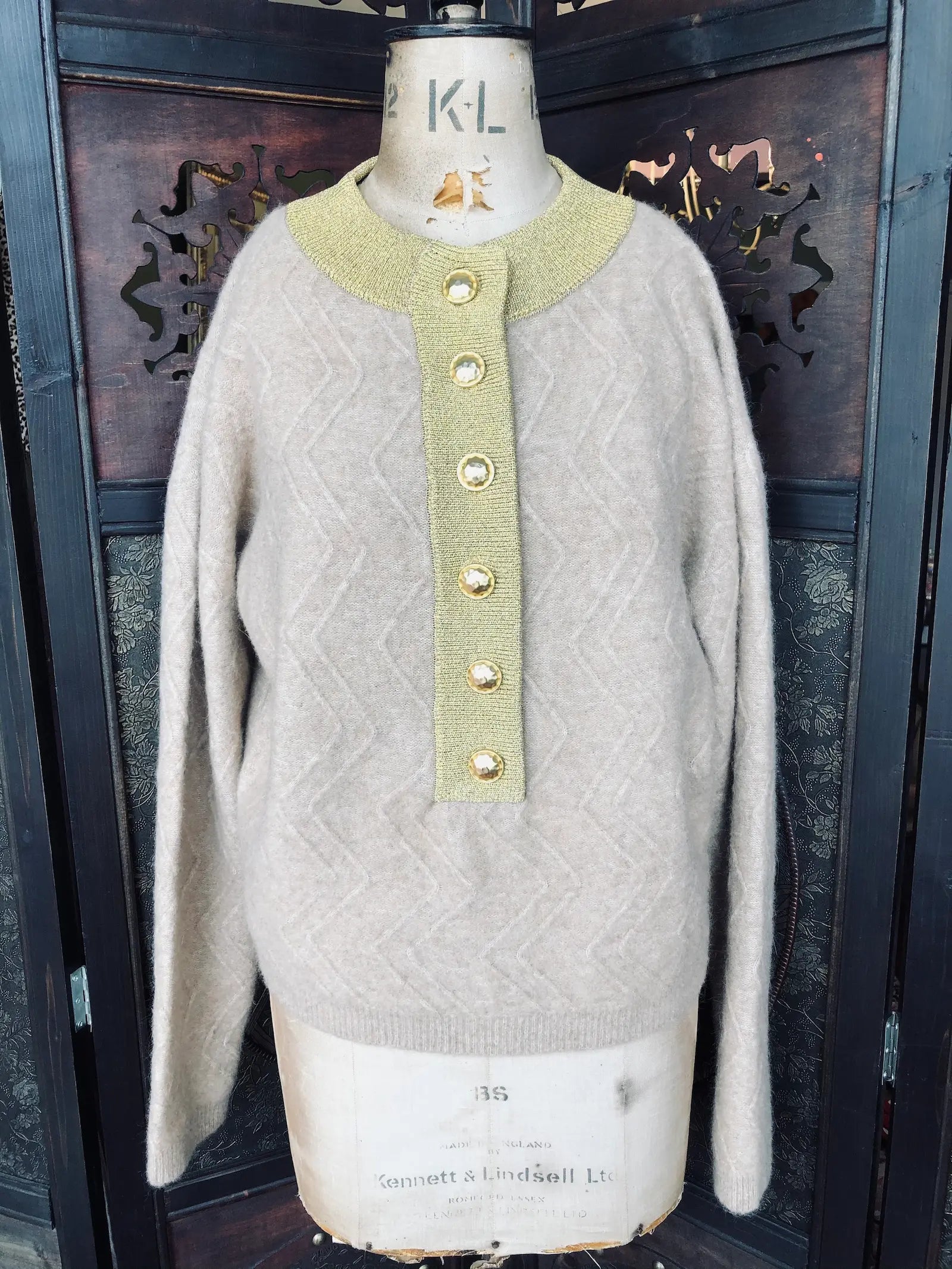 Broochella The Stylist's Closet Michonet beige and gold wool sweater, with gold buttons down the centre front. It is pictured on avintage mannequin. The background is an ornate, dark wood room divider. 