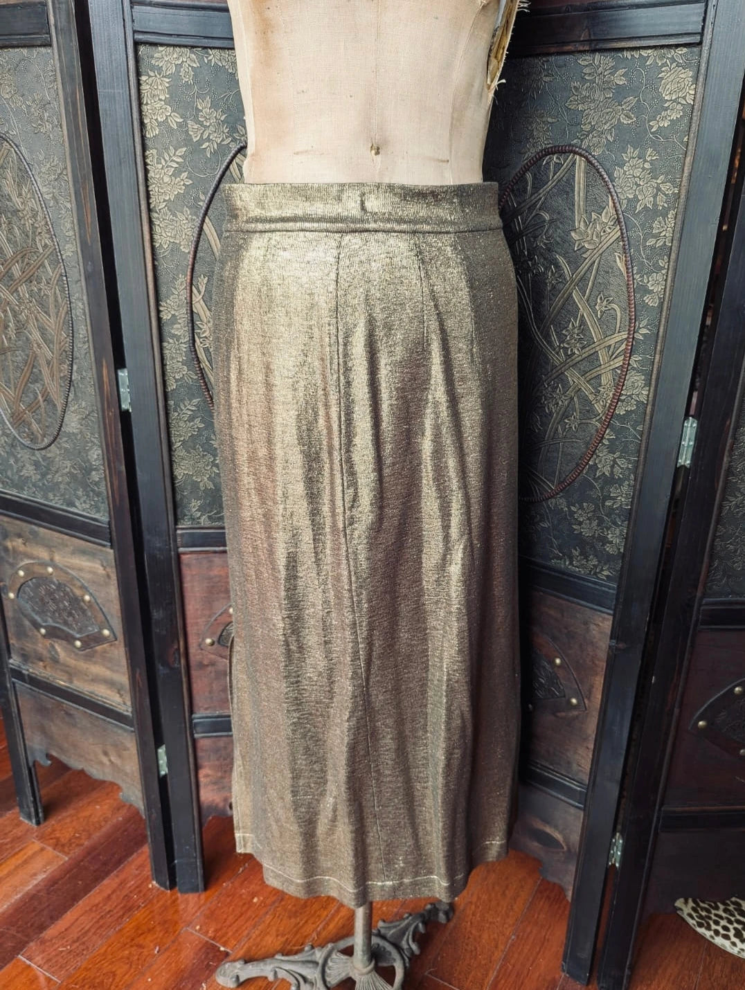 Broochella The Stylist's Closet Michonet designer, metallic gold midi skirt in a size small. It is pictured on a vintage mannequin. The background is an ornate dark wood room divider. 