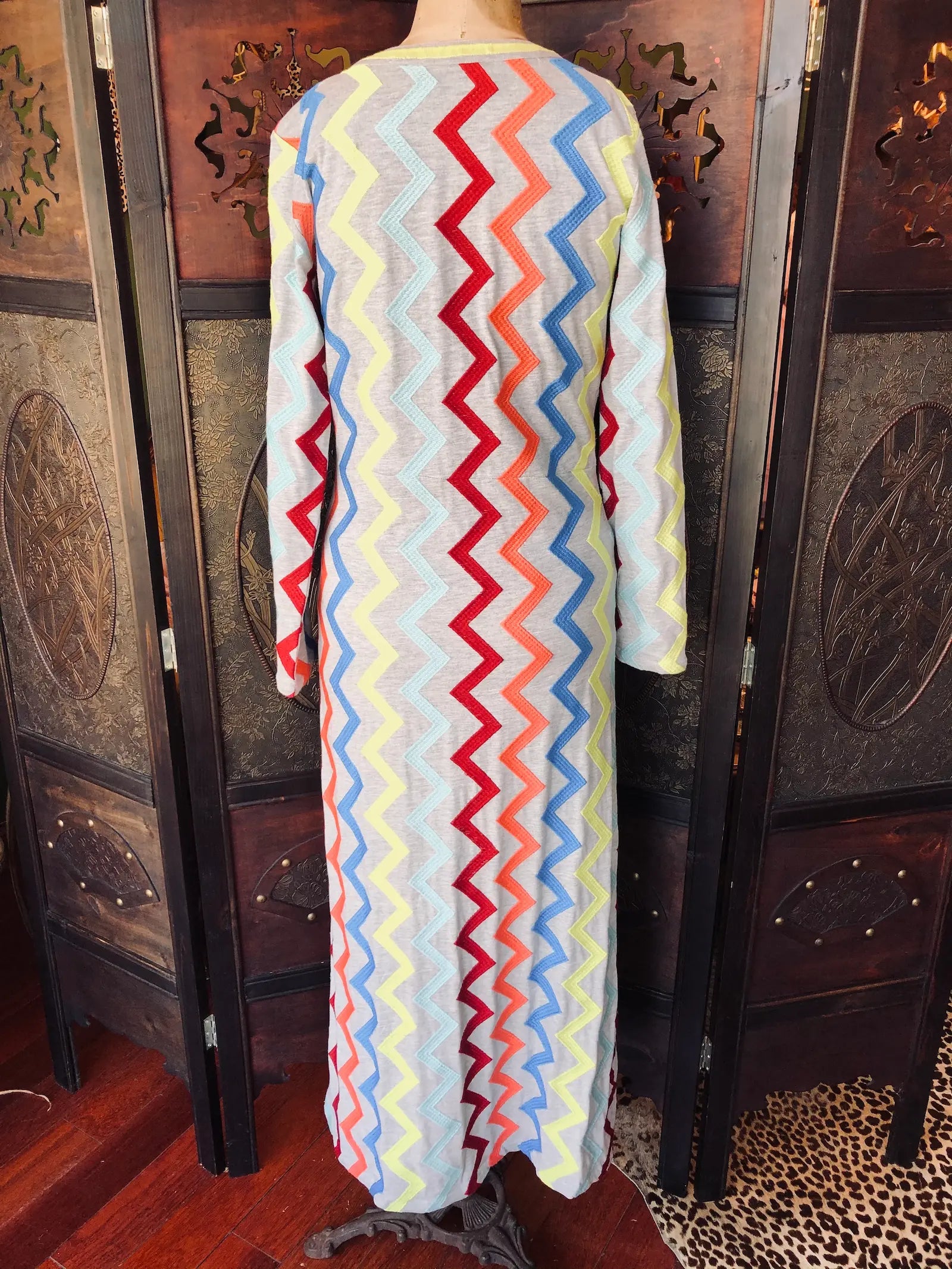 Broochella Michonet size small, grey, long sleeve maxi dress in 100% Cotton. It has long sleeves and multicolour zig-zag embroidery and a V-neckline. It is pictured on a vintage mannequin and is the back view of the dress. The background is an ornate, vintage, dark wood room divider. 