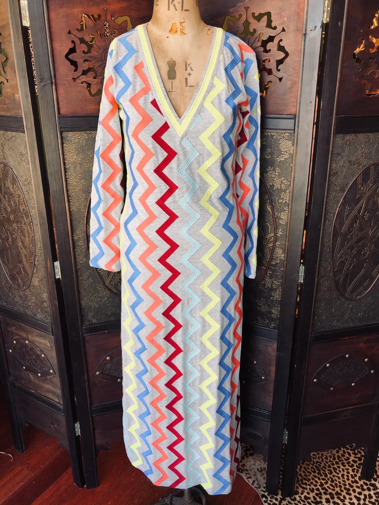 Broochella Michonet size small, grey, long sleeve maxi dress in 100% Cotton. It has long sleeves and multicolour zig-zag embroidery and a V-neckline. It is pictured on a vintage mannequin. The background is an ornate, vintage, dark wood room divider. 